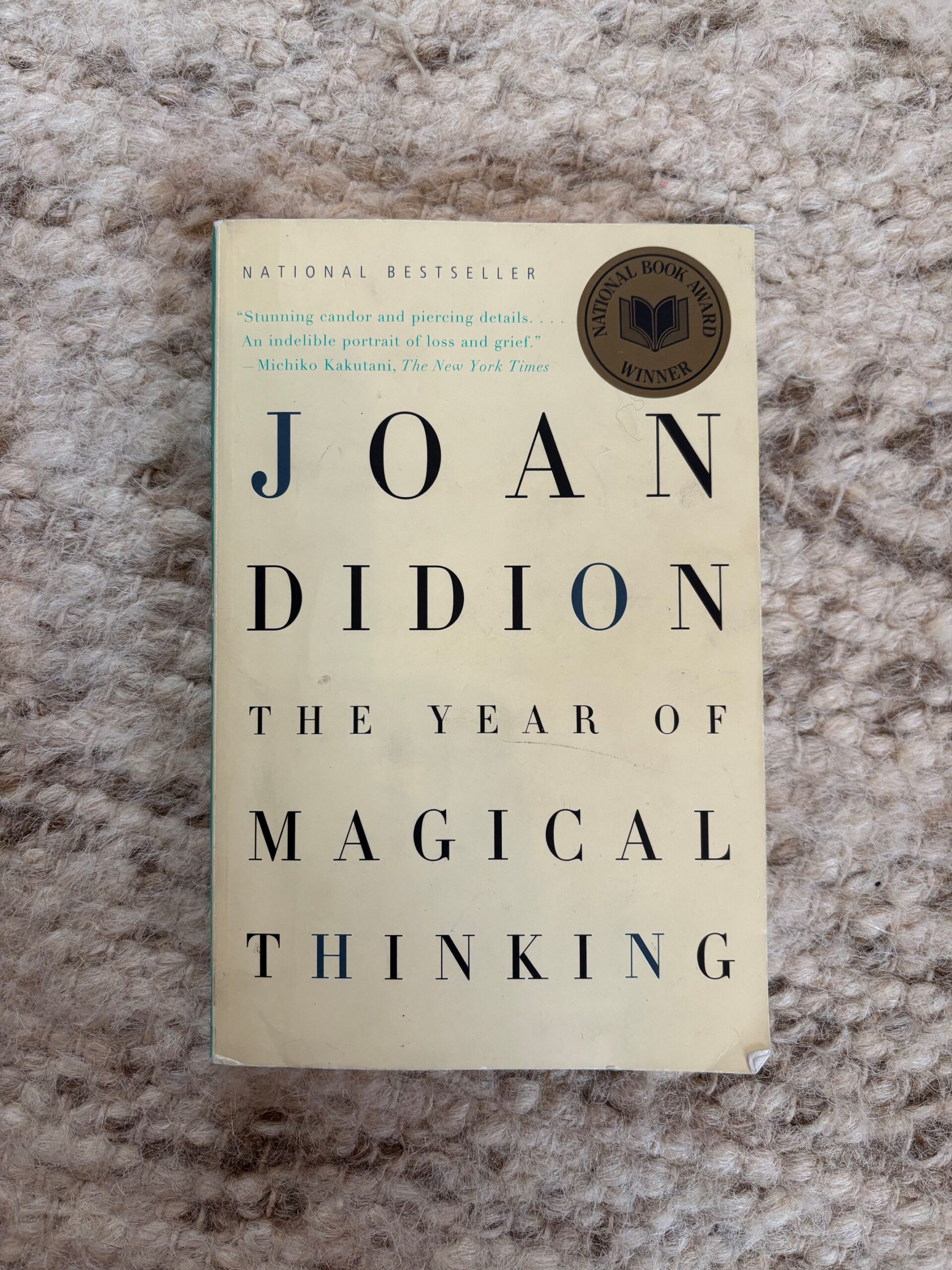 A paperback copy of Joan Didion's The Year of Magical Thinking photographed on a cream-colored wool rug.