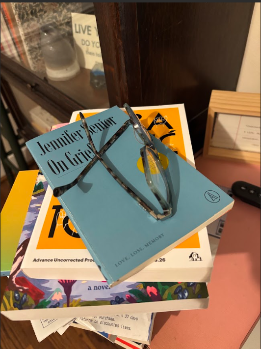On Grief by Jennifer Senior, a simple blue paperback with black text, sits atop a stack of books on a pink desk.