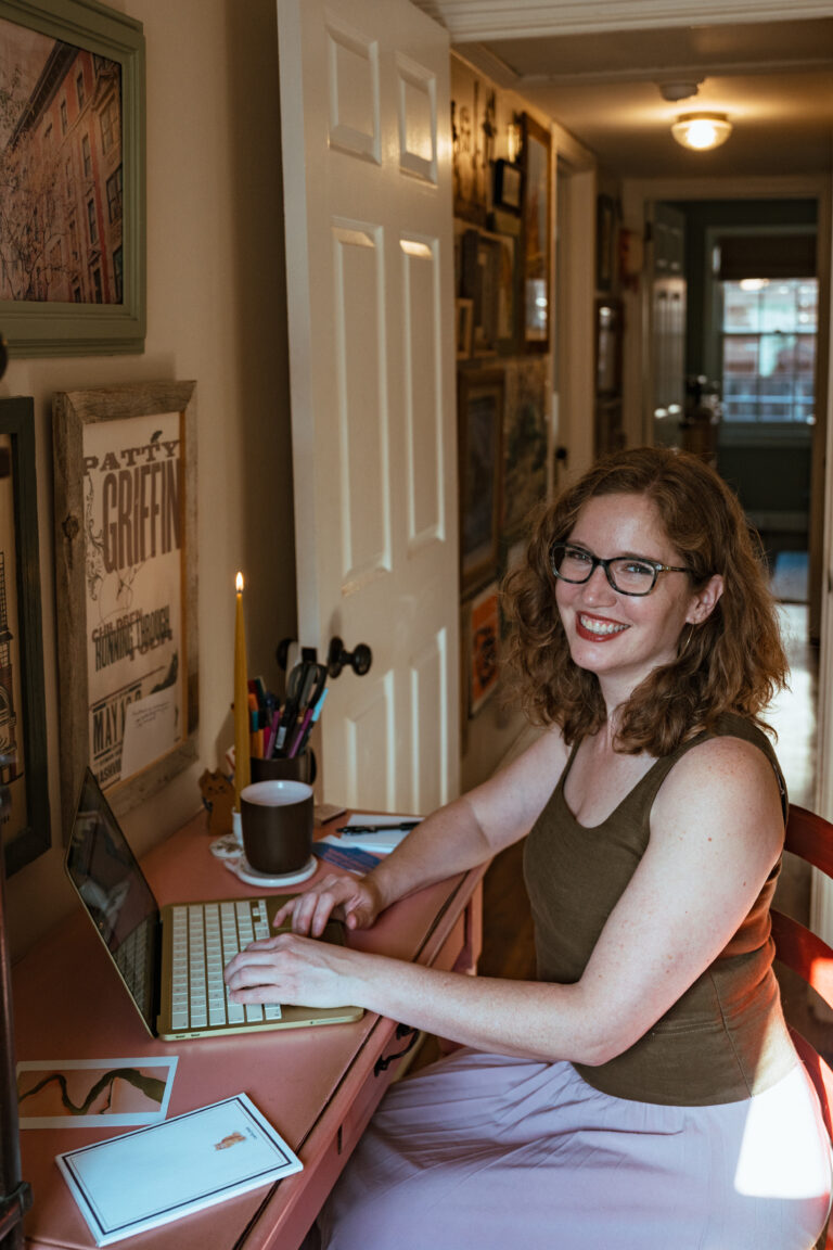 A white woman with coppery, curly hair sits at a pink desk while typing on a citrus Macbook Neo. A beeswax candlestick burns to her right, where a coffee cup also sits. The door to a hallway is open behind her, with artwork hung on the walls.