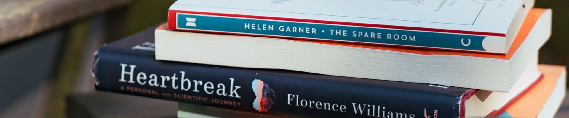 The Spare Room by Helen Garner, a book with its pages facing the camera, Heartbreak by Florence Williams and a book whose title isn't visible sit on an outdoor table in late afternoon.