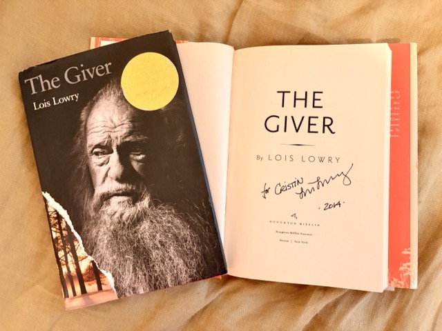 Two editions of The Giver by Lois Lowry are stacked, one open to an autographed title page with the other sitting atop it.