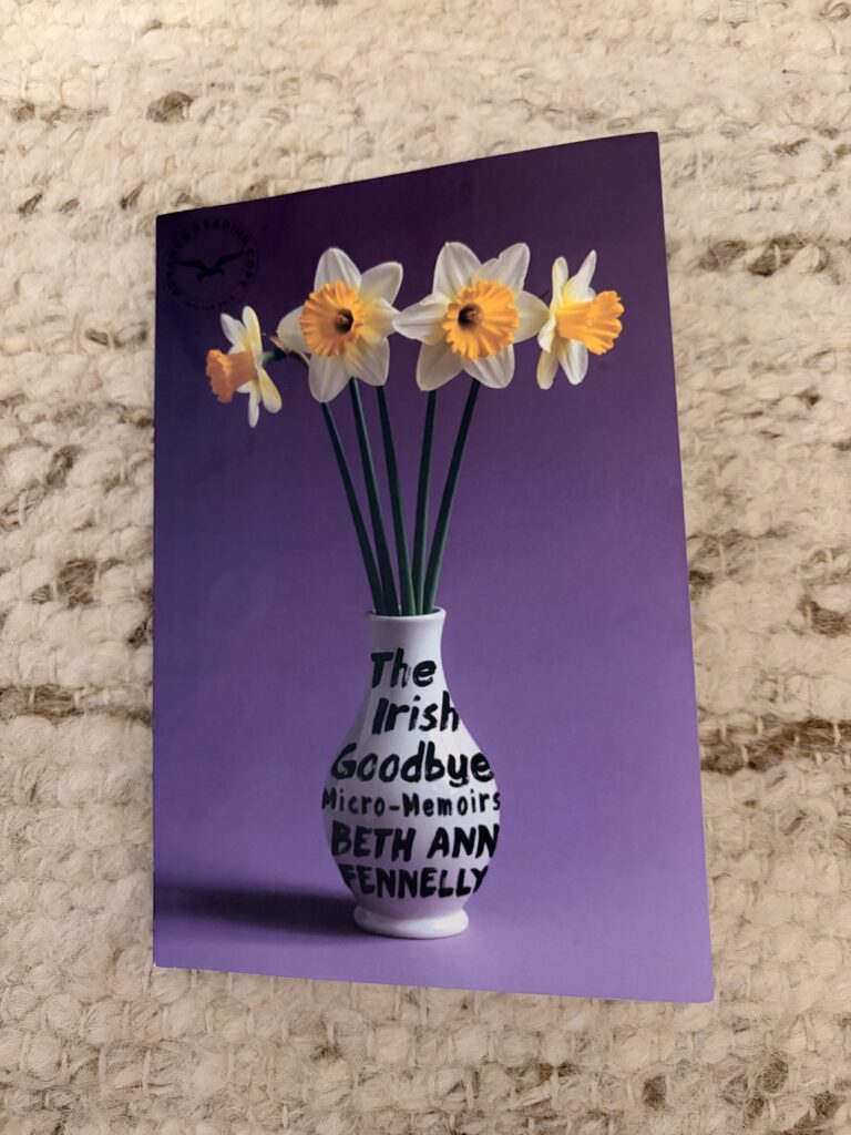 A review copy of The Irish Goodbye: Micro-Memoirs by Beth Ann Fennelly sits on a cream-colored rug. The book's cover is purple and features a vase holding what appear to be faux four daffodils. The book's title and author's name are painted in black on the white vase.